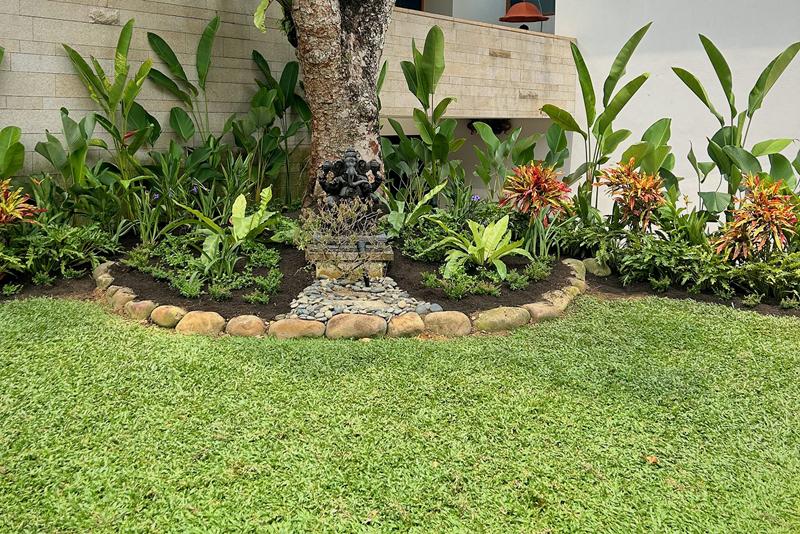 Planting bed around a mature tree with a dark stone statue, river-stone edging and lawn in front.