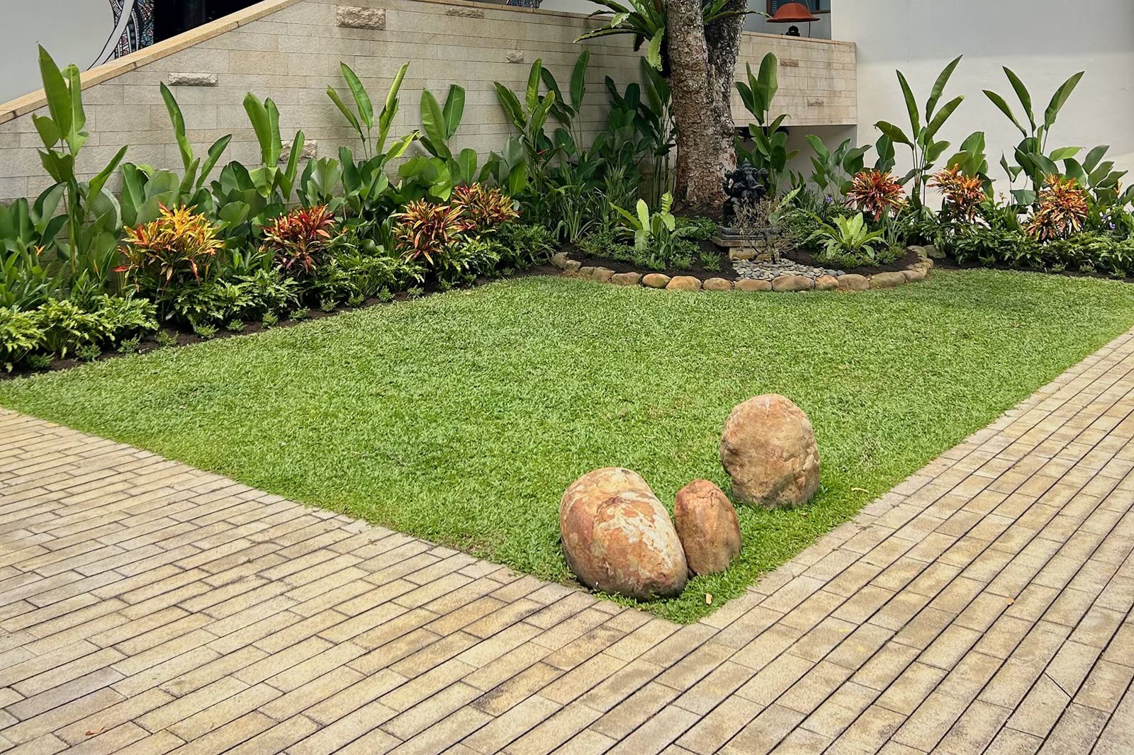 Tropical border of heliconia and crotons along a stone wall, edged in river stones, beside a neat lawn with three boulders.