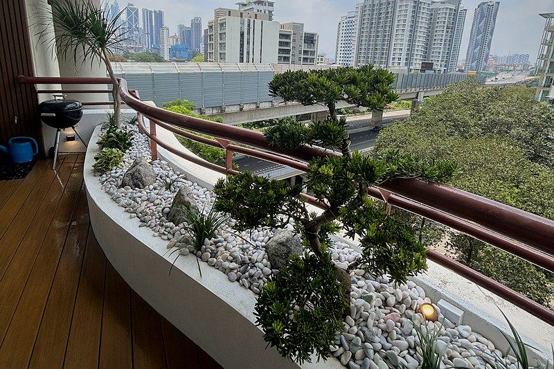 Curved balcony planter with a cloud-pruned tree, white pebbles and granite rocks, city towers beyond the railing.